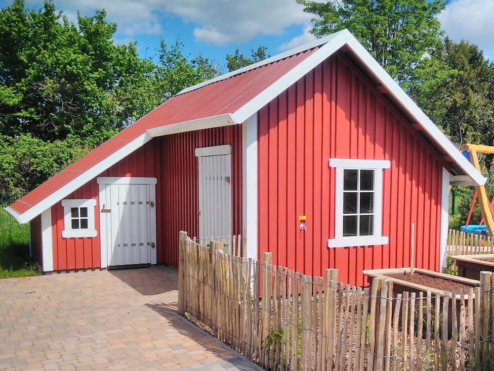 Rotes Holzspielhaus mit wei&szlig;en Fenstern und T&uuml;ren auf naturnahem Au&szlig;engel&auml;nde