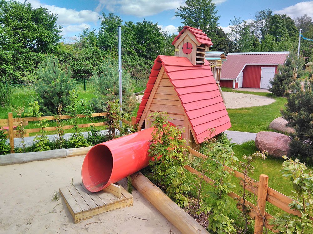 Holzspielhaus mit Röhre – individuelles Spielgerät für Kinder Spielanlage aus Holz mit roter Tunnelrutsche im naturnahen Sandbereich